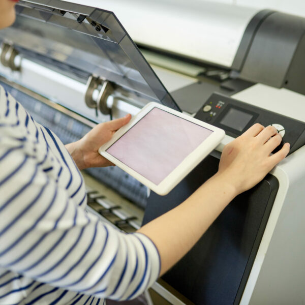 Anonymous young woman using tablet to adjust settings on plotter in printing office.